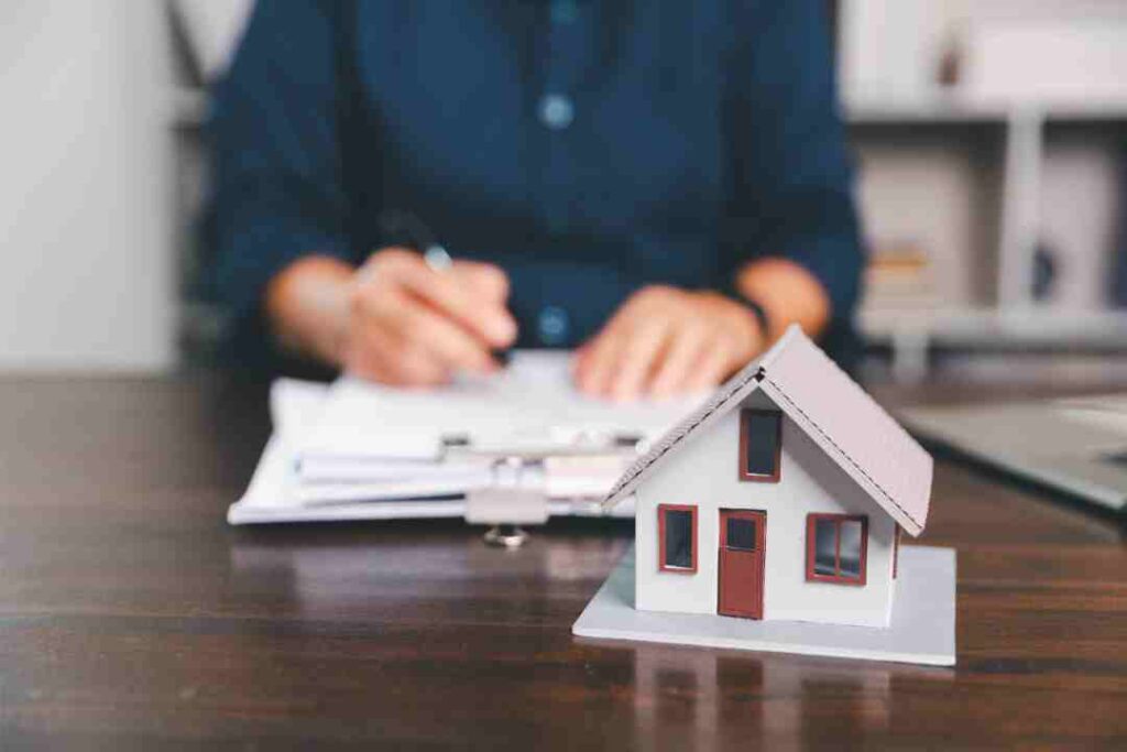 White model house on a desk, person signing documents in the background.